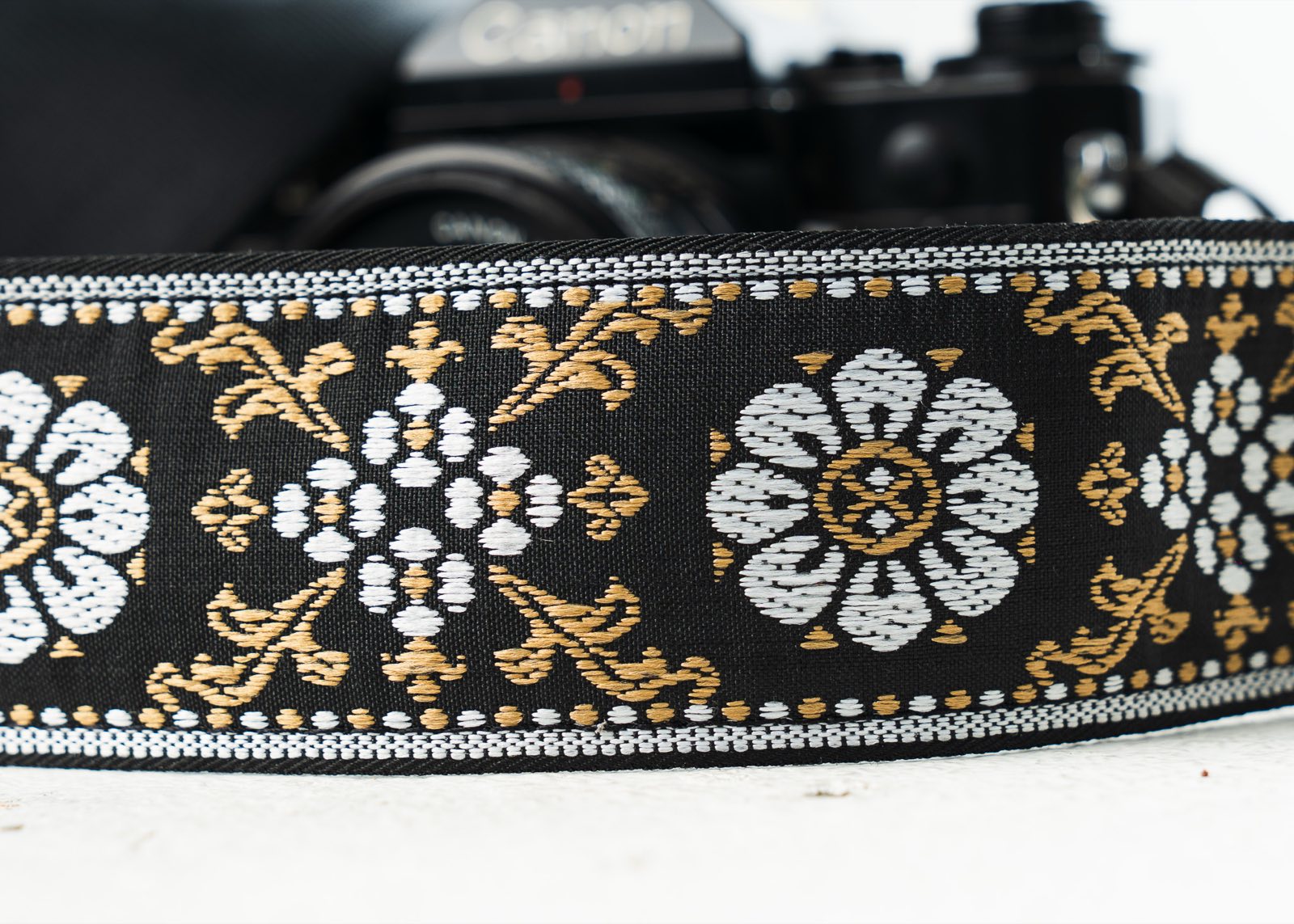 Black, gold and white floral jacquard weave on the camera strap, in front of a vintage Canon